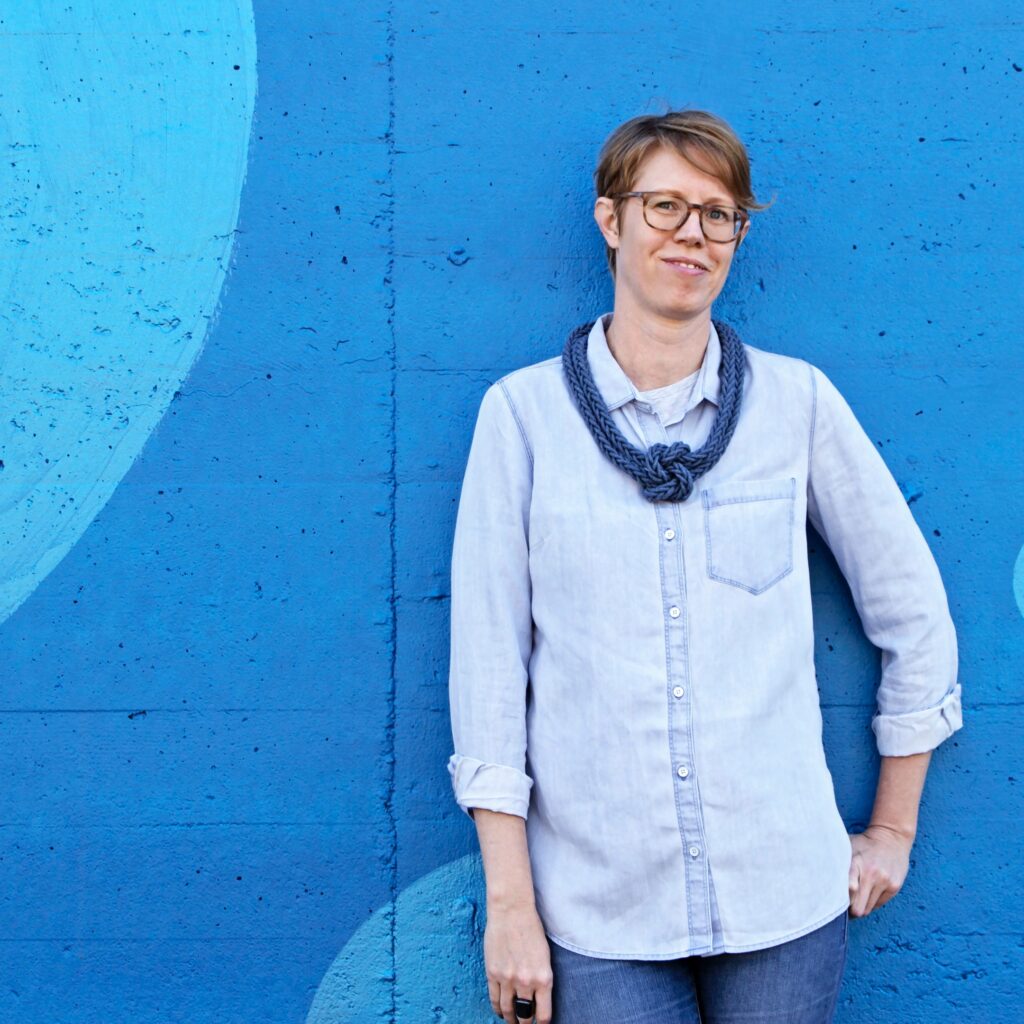 White woman with short red hair and glasses in a chambray shirt against a blue wall