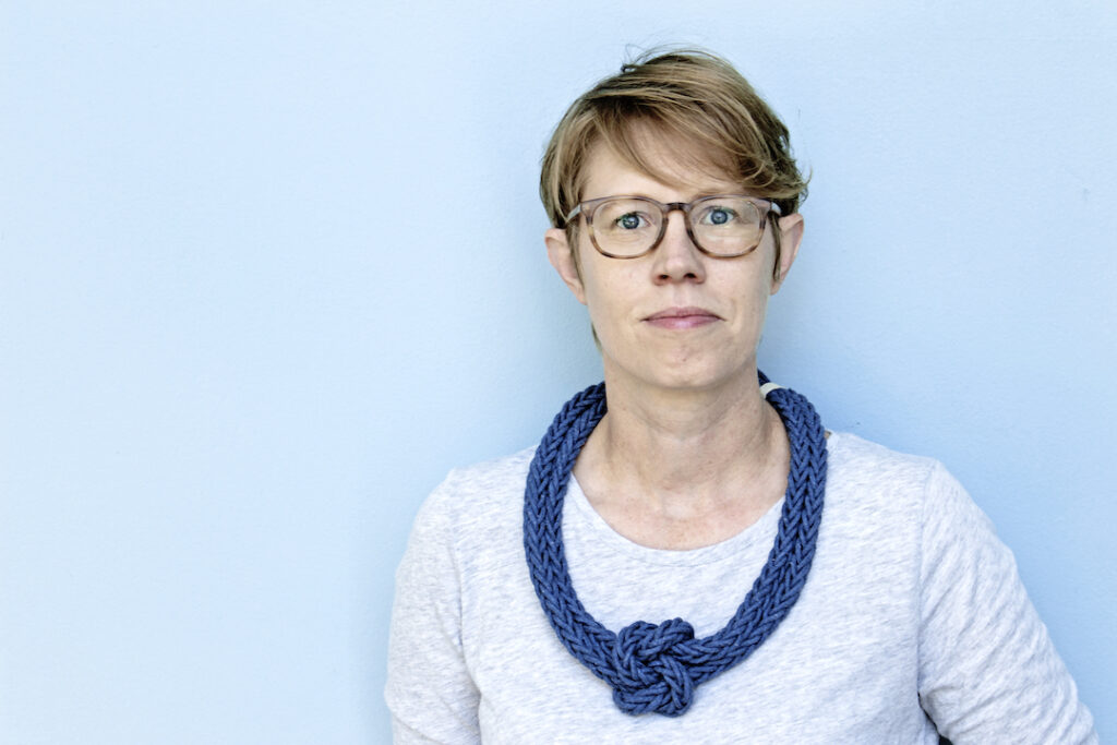 Image of a white woman with short red hair wearing a large blue necklace and a grey shirt with a light blue background